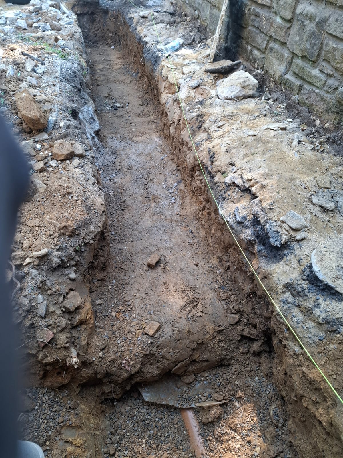 Deep foundation trench excavated alongside an existing stone wall and underground pipe