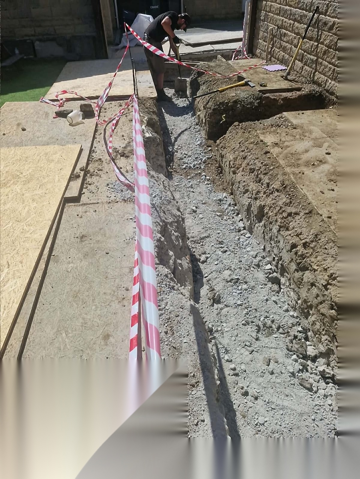 Groundworker digging a trench beside stonework with safety tape protecting the excavation
