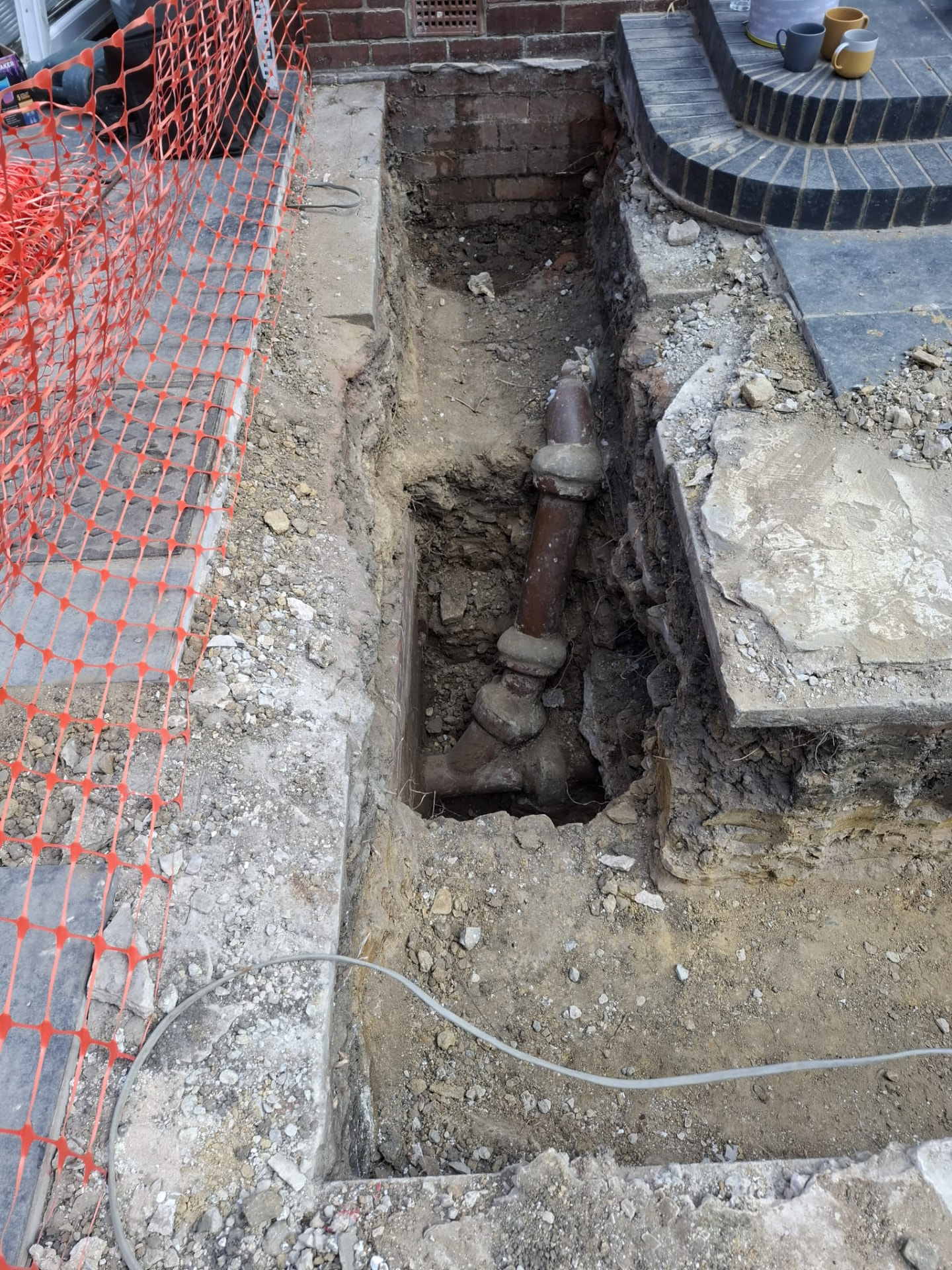 Trench excavation exposing existing underground pipework beside steps, protected by safety fencing