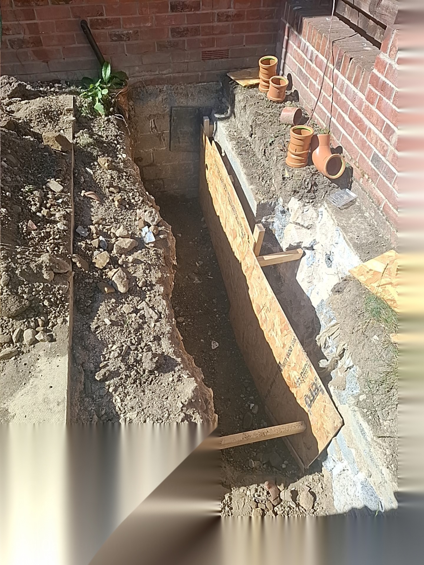 Narrow excavation supported with timber shuttering alongside an existing brick wall